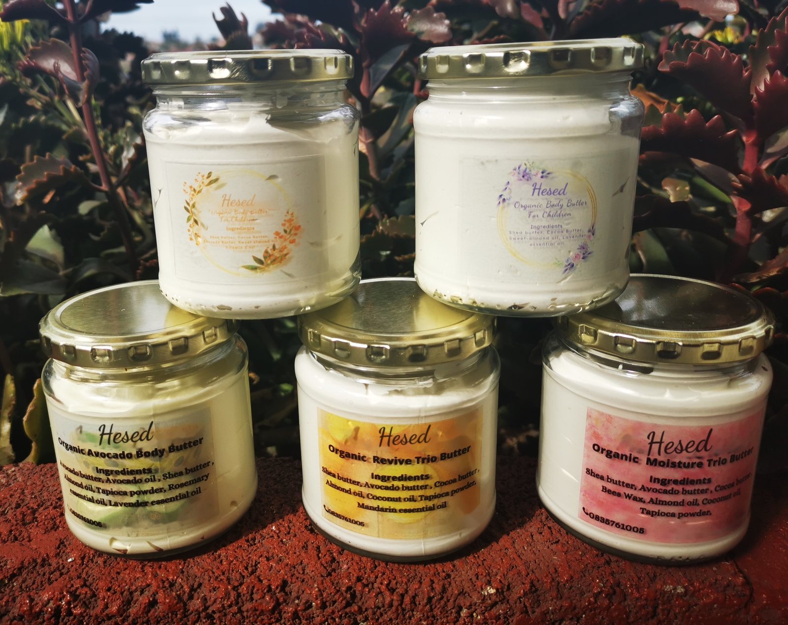 Five Hesed jars stacked on a red brick in the garden: the children's butters on top, and the Avocado, Revive Trio and Moisture Trio butters below